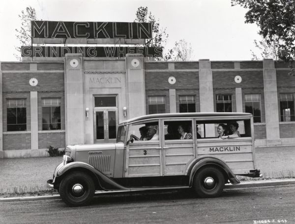 A group of women sit in an International C-1 station wagon parked in front of a building marked: "Macklin Grinding Wheels." A male driver wearing a hat is in the driver's seat. The original caption reads: "Is used to carry office girls to and from work. This C-1 station wagon picks up eight office girls in the morning, takes them home for lunch, brings them back and takes them home at night. Also carries mail."