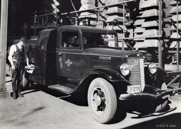 A man stands beside an International C-30 truck owned by the Southern Canada Power Company and places an object into a side compartment. There is a "Danger" sign on the fence behind the truck.