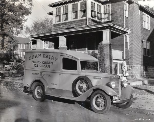 An International truck owned by Dean Dairy is parked along the curb on a street in a residential area. A man is delivering bottles of milk to a house in the background.