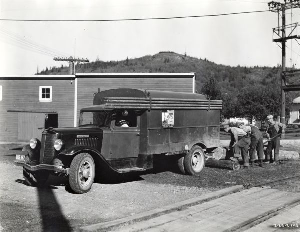 Three men loading pole supports into the back of an International truck owned by the Puget Sound Power and Light Company. In the foreground are railroad tracks, and behind the truck is a low, wooden building and a hill rising above it in the background. The original caption reads: "A C-35 owned by the Puget Sound Power & Light Company, whose headquarters are in Seattle, but this truck is operating out of this firm's branch at Burlington, Washington.  It is used for general maintenance work on the company's many miles of power and light lines in that section of the northwest. The truck is equipped with a complete assortment of repair material to meet any emergency and is kept available for such service day and night. I-36-Z shows it taking on a load of pole supports, these being hauled into the interior of the truck with its own winch powered by the truck engine..."