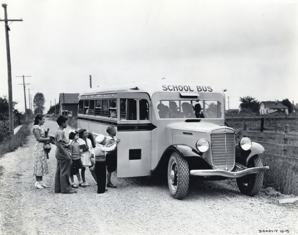 An International Model C-60 school bus is parked on a gravel road in a rural area while children form a line to board. The bus was owned by the schools of Fife, Washington, whose fleet also includes an A model and two C-40's.
