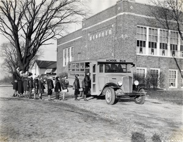 Children wearing winter coats and hats forming a line to get on a school bus parked in the drive in front of what appears to be a school building. The text on the side of the bus reads, "Springfield...School."