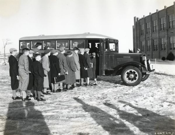 Boys wearing winter coats and hats are lining up in the snow to board an International school bus parked in front of Hershey Industrial School. All the boys are wearing calf-length coats or short pants (knickers), patterned socks and dress shoes.