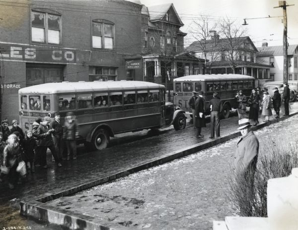 Two International buses owned by Monongalia County Schools parked in a street. Children and adults stand on the sidewalks surrounding the buses. A commercial building and homes are in the background.