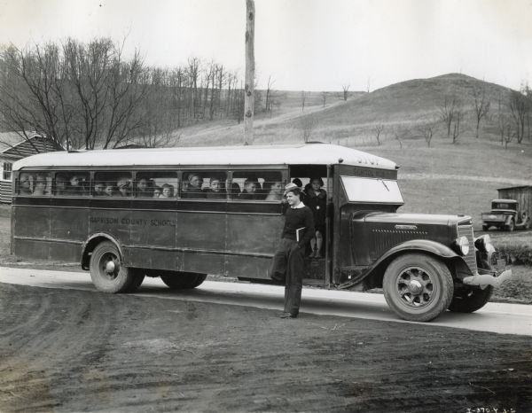 A boy exits an International bus wearing a hat and carrying a book, and a young girl stands on the steps behind him. Other children look out the window from their seats. The bus is marked: "Harrison County Schools" and it is parked alongside a road in a rural setting, with a house in the background, and a truck parked near a wooden shed.