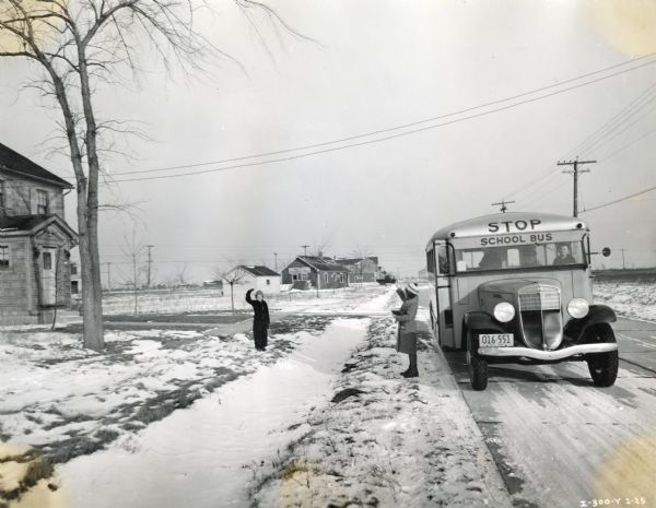 A school bus stops in front of a house in a rural area as two children appear to wave goodbye to each other in the front yard. There is snow on the ground.