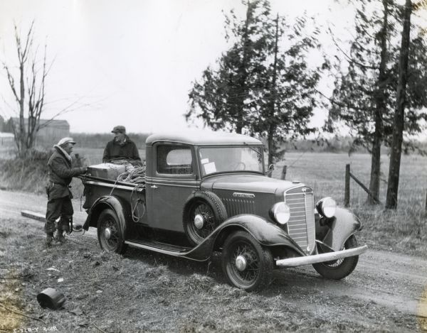 Two men stand near the rear of an International C-1 truck recently delivered to a Puget Sound Light and Power Company branch. Piles of wire and other equipment are loaded in the back of the truck which is parked on the side of a road in a rural area. The original caption reads: "This unit was immediately pressed into service to assist in restoring light and power service that had been completely disrupted in the northern part of this state by one of the worst blizzards and silver thaws that had been experienced in many years."