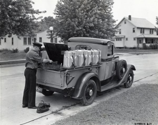 A man in a work uniform and hat is placing a wooden tray of tools into the back of an International pickup truck. The truck is parked along the side of a residential road and its passenger door is marked: "Springfield Gas."