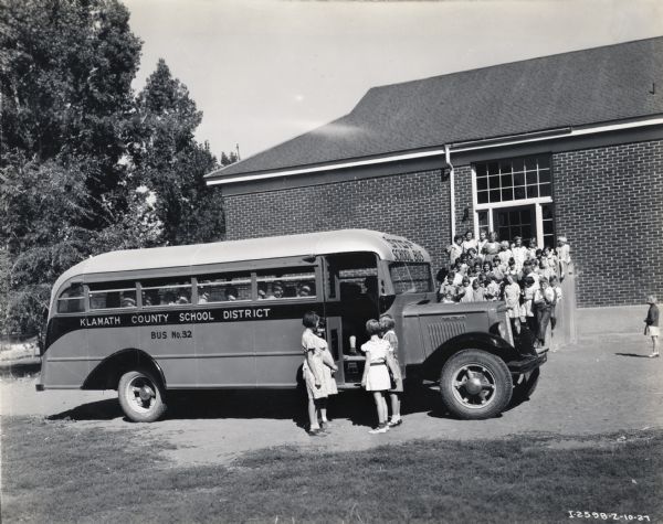 Children stand near the open door of an International school bus marked "Klamath County School District, Bus No.32" while others sit on the seats inside. The bus is parked in front of a school building and a large group of children stand on the front steps. The original caption reads: "Two C-30 school buses operated by Klamath County, Oregon, equipped with Pacific steel bodies shown at Henley Grade School. Martin Bridges, Midland, Oregon, and A.L. Wilkinson, Route 1, box 135, Klamath Falls, Oregon, drivers: A.N. Reeder, Route 2, Box 177, Klamath Falls, Oregon, driver and salesman; Fred Peterson, Courthouse, Klamath Falls, Oregon, school official."