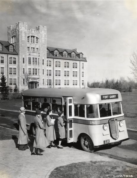 Elevated view of men and women lined up on sidewalk to enter an International bus owned by Winnipeg Electric Company. The bus is marked: "Fort Garry." A large stone building and lawn are in the background.