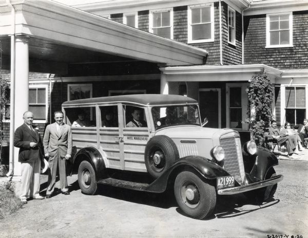 Two men stand beside an International C-1 station wagon ("woody") owned by Hotel Marblehead. Men and women passengers sit inside the vehicle along with the driver, and three individuals sit on lawn chairs in the sun beside the entrance to the hotel.