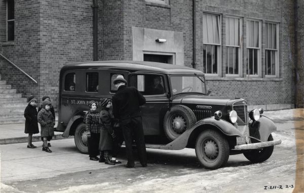 A man helps children get into an International C-1 school bus (station wagon). In the background is a large brick building.