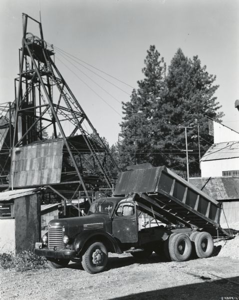 An International KB-8-F dump truck owned by the Empire-Star Mines Company, Limited, parked in front of several buildings with its bed raised to ore(?). A man is standing on the running board on the driver's side to watch the dumping. The original caption reads: "There is a lot of gold-mining history dating back to the turbulent days of 1854 depicted in the background of this picture of an International KB-8-F dump truck owned by the Empire-Star Mines Company, Limited, of Grass Valley, California. It shows a head frame 116 feet high, located directly over the company's central shaft. This shaft is vertical and 3650 feet deep. Two miles away is the company's Empire Shaft, and there is also the Pennsylvania shaft. Operations are now conducted on a limited scale because of Government restrictions on gold mining decree, and so the ore taken from the Central shaft shown is transported by motor truck to the mill at the Empire shaft instead of the mill at the Central shaft. Besides the truck shown an International K-6 is used to haul the ore to the other stamp mill two miles away. Each truck makes about 25 round trips a day."