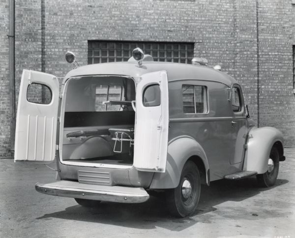 Rear view of an International KB-2 panel truck converted into an ambulance. The rear doors are open, allowing a view of the collapsible bed inside. The original caption reads: "These trucks equipped with Baumgardner cots, overhead stretcher, medicine cabinet, and ventilating unit. These trucks were made for shipment to Siam."