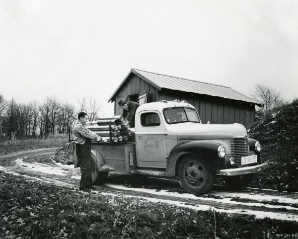 Two men load torpedoes onto the bed of an International truck marked: "The Independent Eastern Torpedo Co." A sign reading: "Explosives" is on the grille of the truck, and a small shed is in the background.