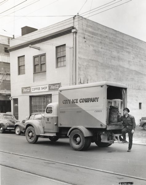 A man removes a bag from the back of an International truck owned by City Ice Company. The truck is parked on a street in front of a coffee shop which advertises Coca-Cola. The original caption reads: "The City Ice Company of Portland, Oregon, which is affiliated with the Northwest Ice & Cold Storage Company, recently purchased four K-5 and one K-8 Internationals. Three of the K-5's and the K-8 are shown at the ice plant in accompanying illustrations. Each is equipped with van-panel body which is attractively finished in blue and yellow. The small K-5's are operated on retail routes with an average of 235 stops and 42 miles per day. The larger K-8 delivers ice mainly to re-sale boxes located in various parts of Portland and suburbs within a radius of 8 miles of the plant. The City Ice Company has been buying Internationals since 1930. Its fleet now consists of 15 units."