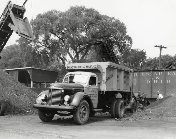 Front three-quarter view of an International KB-7 truck used to deliver coal by Evanston Fuel and Material Company. The truck is parked in front of railroad cars as coal is loaded onto its bed.