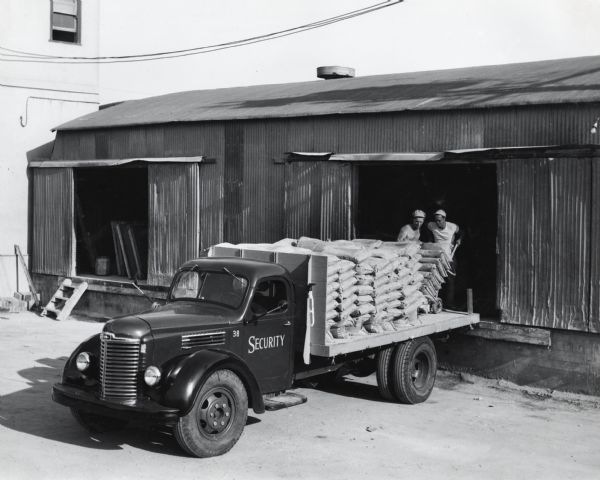 Slightly elevated view of two men using a dolly from inside a shed to load bags of cement onto the bed of an International KBS-7 truck owned by Security Materials Company. The truck is hauling approximately 192 sacks of cement. The original caption reads: "An International KBS-7 with 176-inch wheelbase owned by the Security Materials Company of Los Angeles, California. This truck, one of the nine Internationals owned by the Security Materials Company, is shown hauling approximately 192 sacks of cement."