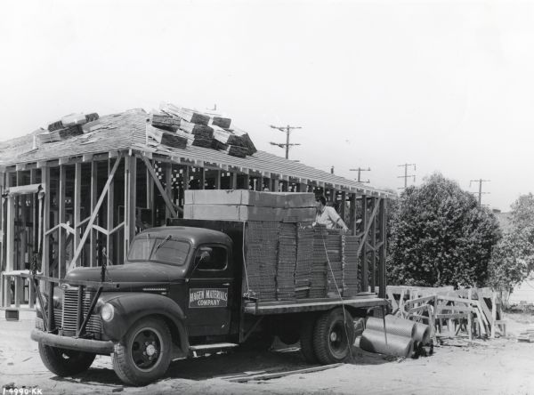 A man stacks building materials on the bed of an International KBS-5 truck with a 159-inch wheelbase owned by the Hagen Materials Company. The truck is parked in front of an unfinished building on a construction site.