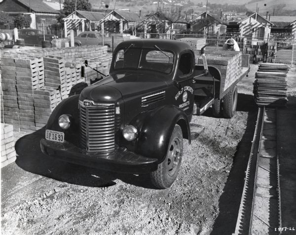 An International Model KB-7 truck used to transport concrete blocks parked on a work or storage site surrounded by building materials. A man loads concrete blocks onto the bed of the truck. In the background is a fence, and on the other side of the street are houses and yards, and hills beyond. The original caption reads, "International Model KB-7, 176-inch wheelbase chassis with special dump body without sides, designed to dump a load of concrete blocks with a minimum of breakage. This unit is powered with a Blue Diamond 269 engine, has a 5-speed transmission and a 2-speed 6.14 - 8.25 to 1 rear axle. 9.00 - 20 tires. Unit is owned by the Hurst Concrete Products, Santa Barbara, California. Unit was placed in service November 14, 1947."