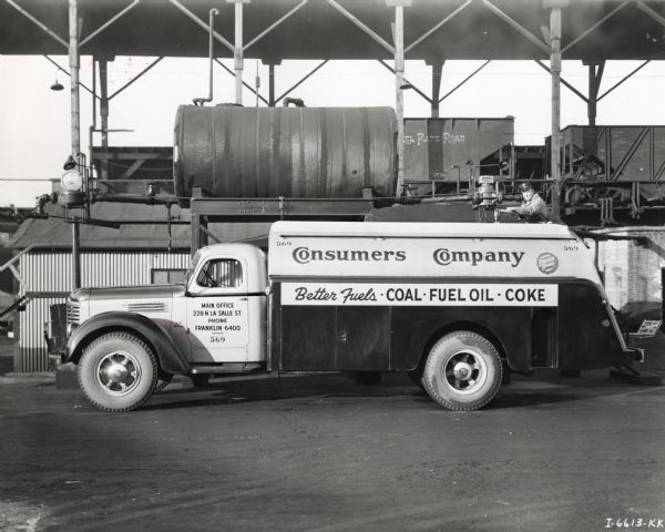 Left side view of an International Model KB-10 truck owned by Consumers Company and outfitted with a fuel oil tank. The text on the truck reads: "Better Fuels - Coal - Fuel Oil - Coke." A man can be seen just above the truck roof as it is being filled from a large tank. In the background are railroad cars on an elevated track under a large roof.