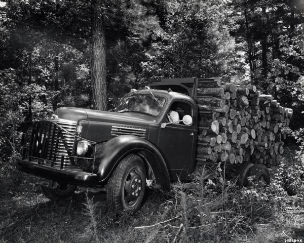 Three-quarter side view of a man driving an International KB-5 Pulpwood Special heavily loaded with logs through a wooded area. The truck was owned by Johns-Manville Corporation and featured a 158-inch wheelbase.