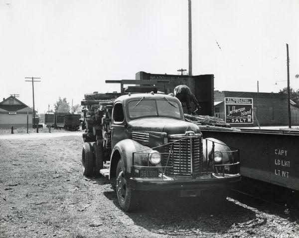 A man uses an International KB-5 Pulpwood Special to haul logs. The truck is parked in a dirt lot next to what appears to be an open railroad car. There are commercial buildings in the background. A sign on a building on the left says: "Roxie."