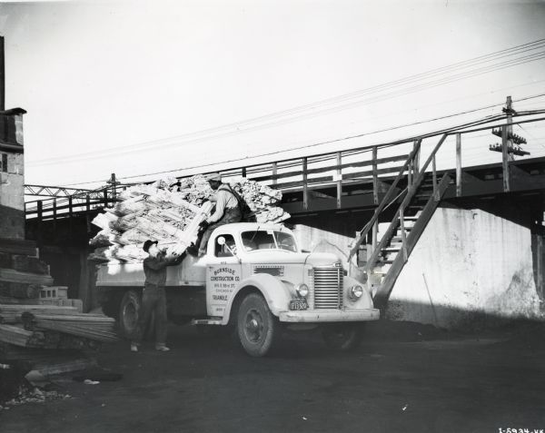 Two men load bundles of plywood(?) onto the bed of an International KB-7 owned by Burnside Construction Company. The truck had a 157-inch wheelbase and a Blue Diamond engine.