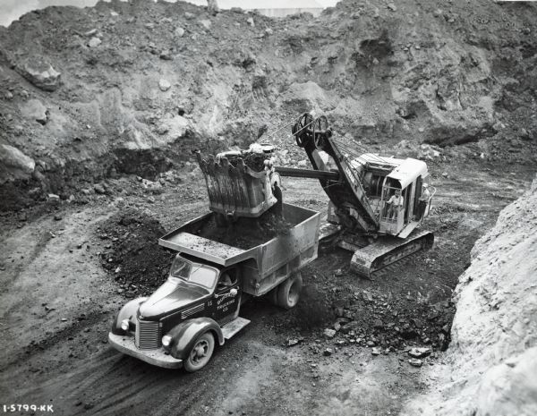 A man loads an International K-7 dump truck with an excavator in a pit. One man is in the driver's seat of the truck, and two men are on the excavator. The original caption reads: "K-7, 146-inch wheelbase. BLD 269 engine. F-51-C transmission. Straight single-reduction rear axle 7.16-1. Penn body and hoist. 8-yard water level body capacity hauling about 8 tons under same conditions as I-5806-KK."