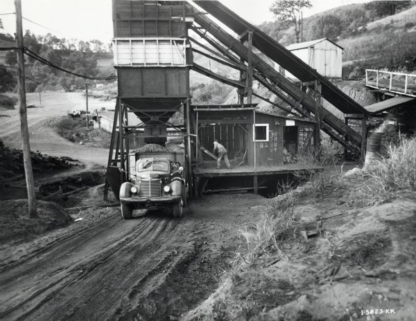 Man in a shed releases coal from a chute into the back of an International KB-11-F truck owned by the Coal Service Corporation. Another man is in the driver's seat of the truck with the door partially open. Other outbuildings are behind the shed and chute, with steep hills and trees surrounding the area.