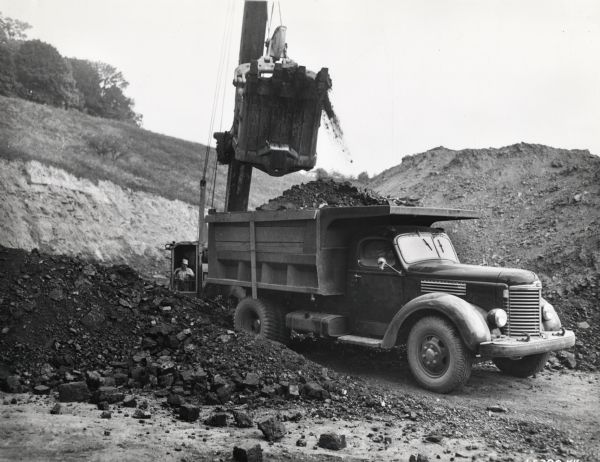 Man uses an excavator to fill an International KBS-7 dump truck with coal. Behind the excavator is a steep hill with trees on top. The coal is being handled by Paul Haul of Bridgeport, West Virginia, the contract hauler for Indian Coal Company of Taylor County, West Virginia. The specifications for the KBS-7 include a 146-inch wheelbase, BLD-269 engine, F-51-C transmission, Eaton 2-speed rear axle, Penn body and hoist, a capacity of 10 yards and an average haul of 9 tons over a 7-mile round trip from the loading zone with shovel to railroad tipple, and about 4 miles of dirt road operation.