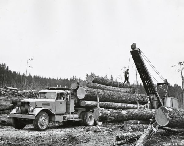 Men using a crane to load logs onto the trailer of an International W-6564-OH truck. A man is swinging from the crane's cable over the logs on the truck. Another man is in the driver's seat of the truck.