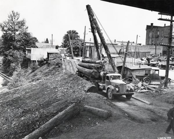 An International K-model truck with logs piled on its trailer parked next to a dirt ramp or embankment leading to a river(?). Industrial buildings are to the right. The truck was used in logging from Santiam Country of Oregon to the dumps on the Wilamette River at Albany, Oregon. The sign on the building in the background reads: "Smith's Glass Service; Auto-Window-Plate-Mirrors."