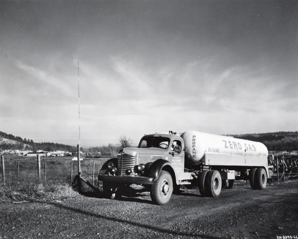 A man sits behind the wheel of an International KB-11 tanker truck parked alongside a gravel road near a fence. The truck was owned by Butane Wholesale Gas Company of Little Rock, Arkansas. In the background are fields and houses surrounded by trees and hills.