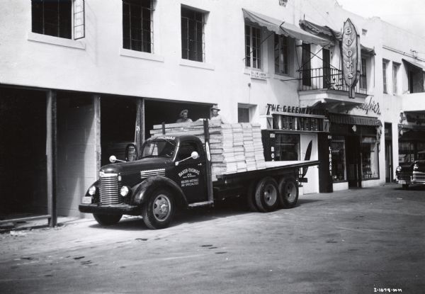 An International Model KB-6-F truck used by Baker-Thomas Company parked near the front of a library building. Two men stand on the bed of the truck with stacks of plywood(?).
