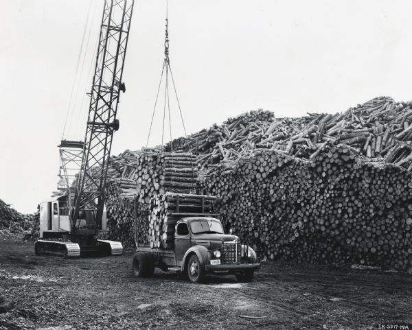 A man uses a crane to load a pallet of logs onto the bed of an International KBS-6 pulpwood special truck. The crane and truck are parked in front of a large pile of logs. The original caption reads: "The International KBS-6 pulpwood special motor truck is doing a most satisfactory job of hauling in the tough conditions that prevail in this type of work for two owners in the vicinity of Brunswick, Georgia. One of the trucks owned by W.T. Sumler, 1628 Johnson Street, Brunswick, is shown out in the woods, 43 miles south of Brunswick, taking its two loaded pallets. The total load of these two pallets, which are rectangular sleds with a tubular upright at each corner, is two and one-half pulpwood units or about three cords. Mr. Sumler is shown in one of the illustrations supervising the winching of loaded pallets up on the truck over two sled runners fastened to the rear of the truck. The other International KBS-6 pulpwood special motor truck, which is owned by George Sears of Hortense, Georgia, is shown also with a load of two pallets or two an done-half units...plant of the Brunswick Pulp & Paper Company."