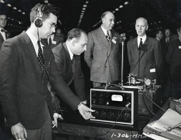 Two men are speaking into a microphone labeled "WGL", while a man in the foreground on the left is wearing headphones and operating radio equipment. Men are watching from behind, and other men are mingling in what appears to be a large building with many rows of lights on the ceiling. The man third from the left may be John L. McCaffrey, president of International Harvester Company.