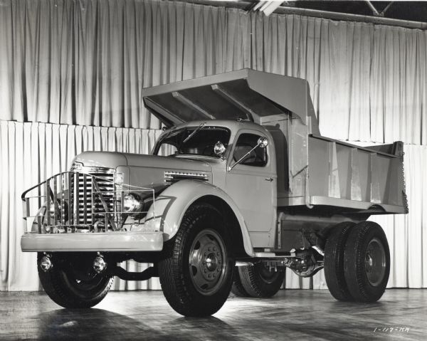 Three-quarter front view of an International KB-11 dump-truck from the driver's side, on display inside a building in front of a hanging backdrop of curtains.