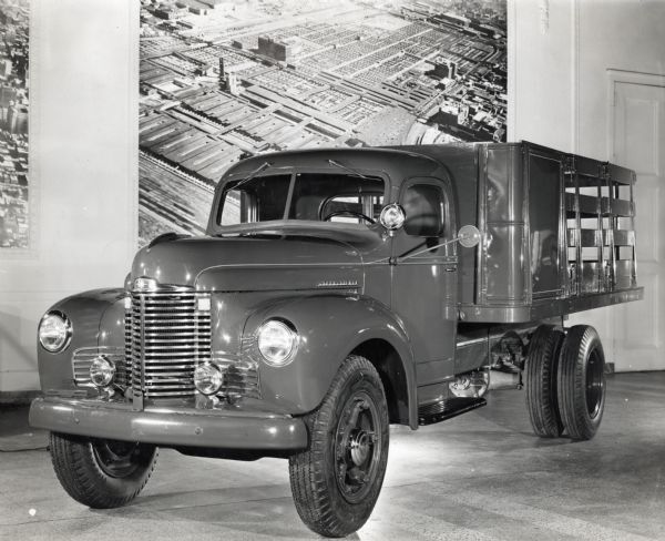 Three-quarter front driver's side view of an International KB-5 truck sitting on display indoors. Mounted on the wall behind the truck are large aerial  photographs of what appears to be buildings, perhaps an International Factory, in an industrial area.