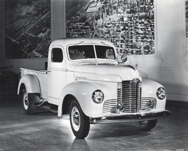 Three-quarter front and passenger's side view of an International KB-1 truck parked in what appears to be a showroom. Hanging on the wall behind the automobile are large aerial photographs of what appear to be industrial buildings, possibly an International factory.