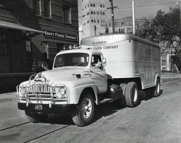 A driver parks an International L-180 truck and Trailmobile trailer owned by the Kraft Foods Company along the side of a road in front of the company's plant. The original caption reads: "The new L-180 International shown at the Kraft Foods Company's plant in San Antonio is one of ten motor trucks operated by the company in San Antonio, eight of which are Internationals. The trailmobile trailer is insulated and 18 feet long. It has double doors at the rear and also a door at one side. It is used to make store to store deliveries and averages 40 stops a day. The wheelbase of the truck is 142 inches. The truck is painted a very attractive attention-getting light yellow and white-shaded blue lettering."