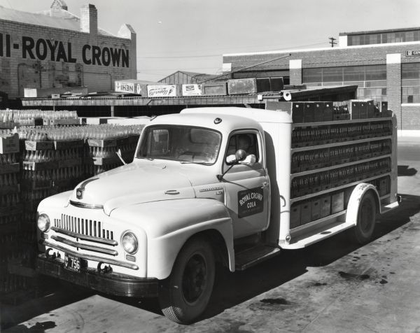 A man parks an International Model L-160 truck owned by the Royal Crown Cola Company near stacks of empty bottles in crates outdoors at the company factory. Boxes of soda bottles are loaded onto the back of the truck. The original caption reads: "A recent purchase of the Nehi-Royal Crown Bottling Company of El Paso, Texas, is a Model L-160 International equipped with drop-frame chassis and special bottlers' body. It is shown at the company's place of business at 1916 Myrtle Avenue. Its wheelbase is 154 inches. The body was made by the P & R Body Company of El Paso. It is 12 feet long, has four decks, and has a total capacity of 235 cases. The company is a partnership consisting of R.R. Ritter, who is shown in one of the illustrations talking to help, Julian Murrufu; an dhis brother, W.P. Ritter, who heads a similar establishment in Phoenix. The El Paso fleet consists of 12 units and deliveries are made as far as Alamogordo, some 100 miles north. Four routes are maintained in El Paso with an average of 40 stops per route."