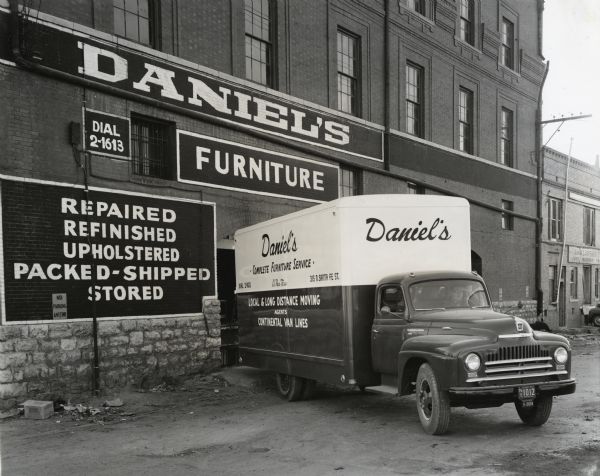 An International L-160 truck owned by the S.L. Daniel Furniture and Mattress Factory, Inc. parks in an alley near what appears to be a company warehouse. A driver backs the truck toward an opened loading door; the text on the side of the truck reads: "Daniel's Complete Furniture Service; Local & Long Distance Moving Agents; Continental Van Line." The original caption reads: "White and rich red are the striking colors that spotlight the new L-160 International furniture van shown as it makes deliveries in various parts of El Paso, Texas. The truck was recently purchased by the S.L. Daniel Furniture and Mattress Factory, Inc. It has a wheelbase of 195 inches. The body is 18 x 8 feet in size, outside dimensions. The concern was founded in 1911 by R.L. Daniels. It was purchased in 1947 by the present organization, of which Mr. M.J. Zimet is president; A.L. Cox, vice president, and H.E. Kramer, treasurer."