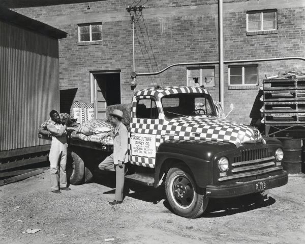 A man unloads a bag of Purina animal feed from the back of a checkered International L-150 truck owned by the Agriculture Supply Company. The truck is parked near a brick building and a shed. Another man stands near its passenger-side door. The original caption reads: "The Agriculture Supply Company of El Paso, dealer for the Ralston Purina Company of St. Louis, has made very effective use of the Purina red-and-white trade-marked checkerboard in the decoration of its new International as shown. The truck, a Model L-150 of 154-inch wheelbase, is equipped with flatbed body. It is one of three trucks used in the delivery of feedstuffs, fertilizers, seed, and insecticides, mostly to farmers within a radius of 30 miles."