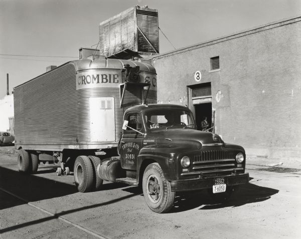 An International L-180 truck owned by Crombie & Company fruit and vegetable dealers parked beside a warehouse in front of a door marked, "3." A woman is standing just inside the door. The original caption reads: "Crombie & Company, wholesale fruit and vegetable dealers of El Paso, Texas, recently purchased the L-180 International shown which brings the total of their fleet to 28 units, 25 of which are Internationals. The truck is shown beside its big El Paso warehouse, the water tower on top of the building being part of a cooling system to control temperatures in the warehouse. This refrigeration is sufficient to care for 45 carloads of produce.  The new L-180 shown has a wheelbase of 145 inches and is one of 14 tractor trucks operated by the concern. Deliveries are made within a radius of 300 miles as far north as Tecumcari and as far east as Midland. Fourteen long distance routes are maintained. The 22-foot Fruehauf trailer shown is of stainless steel with inside width 7-1/2 feet and height 6 feet 6 inches. The concern of Crombie & Company was formed in 1905. W.S. Crombie, Sr. is president, W.S. Crombie, Jr., vice president, and O.C. McConnell, secretary and truck supervisor."