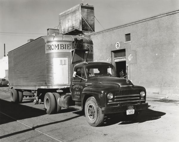 An International L-180 truck owned by Crombie & Company fruit and vegetable dealers parked beside a warehouse in front of a door marked, "3." A woman is standing just inside the door. The original caption reads: "Crombie & Company, wholesale fruit and vegetable dealers of El Paso, Texas, recently purchased the L-180 International shown which brings the total of their fleet to 28 units, 25 of which are Internationals. The truck is shown beside its big El Paso warehouse, the water tower on top of the building being part of a cooling system to control temperatures in the warehouse. This refrigeration is sufficient to care for 45 carloads of produce. 
The new L-180 shown has a wheelbase of 145 inches and is one of 14 tractor trucks operated by the concern. Deliveries are made within a radius of 300 miles as far north as Tecumcari and as far east as Midland. Fourteen long distance routes are maintained. The 22-foot Fruehauf trailer shown is of stainless steel with inside width 7-1/2 feet and height 6 feet 6 inches. The concern of Crombie & Company was formed in 1905. W.S. Crombie, Sr. is president, W.S. Crombie, Jr., vice president, and O.C. McConnell, secretary and truck supervisor."