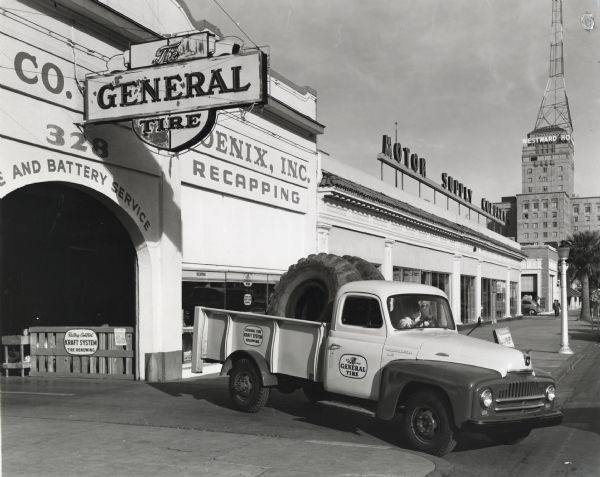A man parks an International L-130 truck owned by the General Tire Company next to the Motor Supply Company building. Two large tires are loaded onto the bed of the truck. A sign reading "Westward Ho" is on a tall building in the background. The original caption reads: "The new L-130 International pickup truck shown in the accompanying illustrations is provided with a special powered tail lift for picking up big earthmoving equipment tires. It is of 124-inch wheelbase with 9-foot pickup body, and is shown in front of the purchaser's place of business, the General Tire Company of Phoenix at 328 North Central Avenue, Phoenix, Arizona. It is shown loaded with two of the big 2100 x 24 tires, each of which weights 800 pounds. The body was made by the Allison Steel Company of Phoenix. The truck is also provided with a compressor for servicing tires out on the job. It travels within a radius of 25 miles."