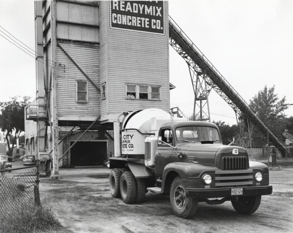 An International LF-190 truck equipped with a concrete mixer in front of a tall building marked: "Readymix Concrete Co." Men are looking out the building's windows. A large conveyer belt is attached to the upper right side of the building, leading down into a fenced area. The original caption reads: "The Twin City Readymix Concrete Company has recently added two new-type Model LF-190's to its all-International fleet. Each of these is of 157-inch wheelbase and equipped with 4-yard mixers. The fleet now consists of 14 units, three of them with Smith mixers and the others with Rex mixers. One of the new LF-190's is shown at the company's plant at 2840 Avenue South, Minneapolis, Minnesota.
