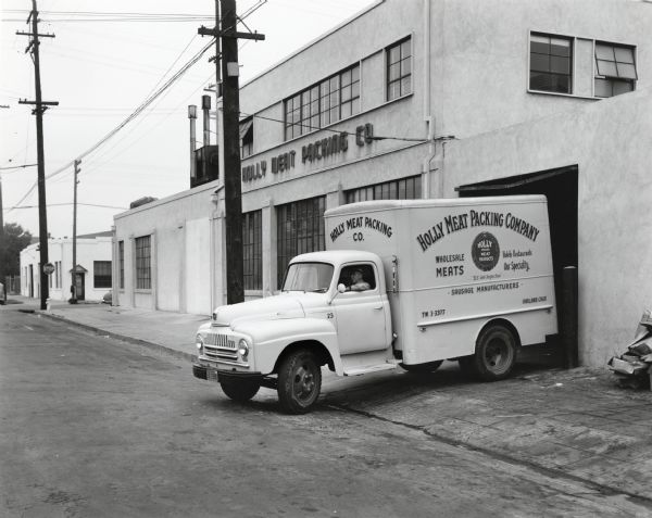 An International L-180 truck owned by the Holly Meat Packing Company is being backed up by a male driver from the street to a loading door at the company's plant. The original caption reads, "...an L-160 with 142-inch wheelbase and a larger L-180 with 154-inch wheelbase and equipped with refrigerated bodies, have just been purchased by the Holly Meat Packing Company. They are shown in front of the company's plant at 2736 Magnolia Street, Oakland, California. The enclosed insulated bodies, 14x7x7 feet and 16x7x7 feet, are painted an attractive canary yellow. The company, a partnership operated by Charles Figone and Louis Figone, is in the wholesale meat and sausage business. Deliveries are made chiefly to restaurants and hotels within a radius of 200 miles of Oakland. Use is made of seven trucks, four of which are Internationals."