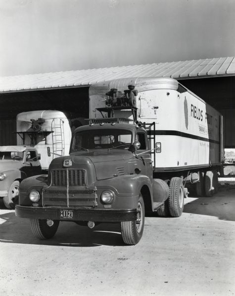 An International L-195 truck owned by the Field Fruit Company parked at an open-air roofed building at a wharf alongside another vehicle. The original caption reads: "The new L-195 International with 30-foot Fruehauf trailer recently purchased by the Field Fruit Company is shown at a wharf in the Port of Brownsville taking on a load of bananas. The bananas were coming direct by a long chain conveyor from a ship which brought the bananas from Southern Mexico and then by another conveyor placed at right angles to the long conveyor to the inside of the big van-type trailer. The Field Fruit Company is located in Memphis in the Texas Panhandle of Northern Texas. The company utilizes two other International transport Internationals (KB-8 and KB-7) beside that shown. They are operated within a radius of 125 miles in Colorado, Oklahoma, Kansas, and Texas. The new L-195 shown has a wheelbase of 133 inches. It makes the long trip to Brownsville two to four times a week. The full load consists of 650 bunches of bananas totaling 25,000 to 30,000 pounds. 
The Port of Brownsville, located just outside the city of Brownsville, is becoming quite an important point for the delivery of bananas from the Isthmus of Tehauntepec. Over 100,000 tons have been delivered the past year and also in 1949. Some 85 percent of these bananas go from the port in motor trucks."